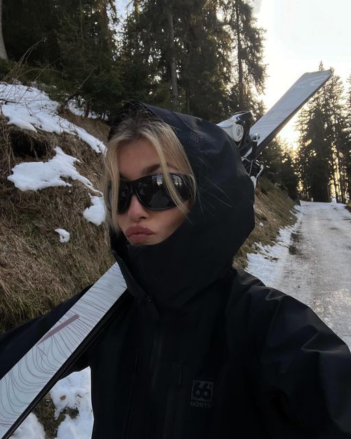 a woman in sunglasses holding a snowboard on the side of a snowy mountain road