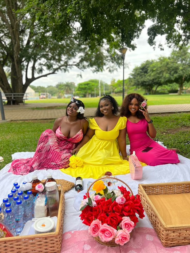 three women sitting on a blanket in the grass with drinks and flowers next to them