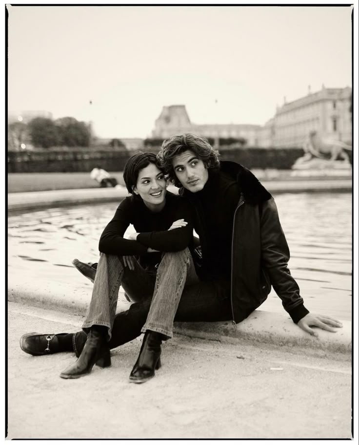 two people sitting next to each other on the ground near water and buildings in the background