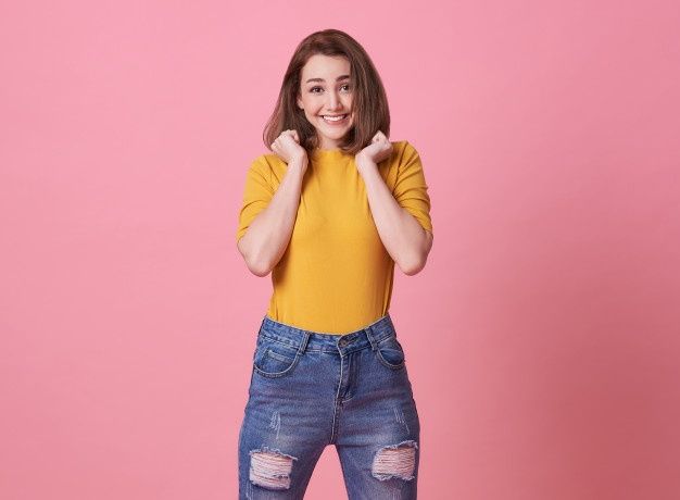 a woman in yellow shirt and ripped jeans posing with her hands behind her head on pink background