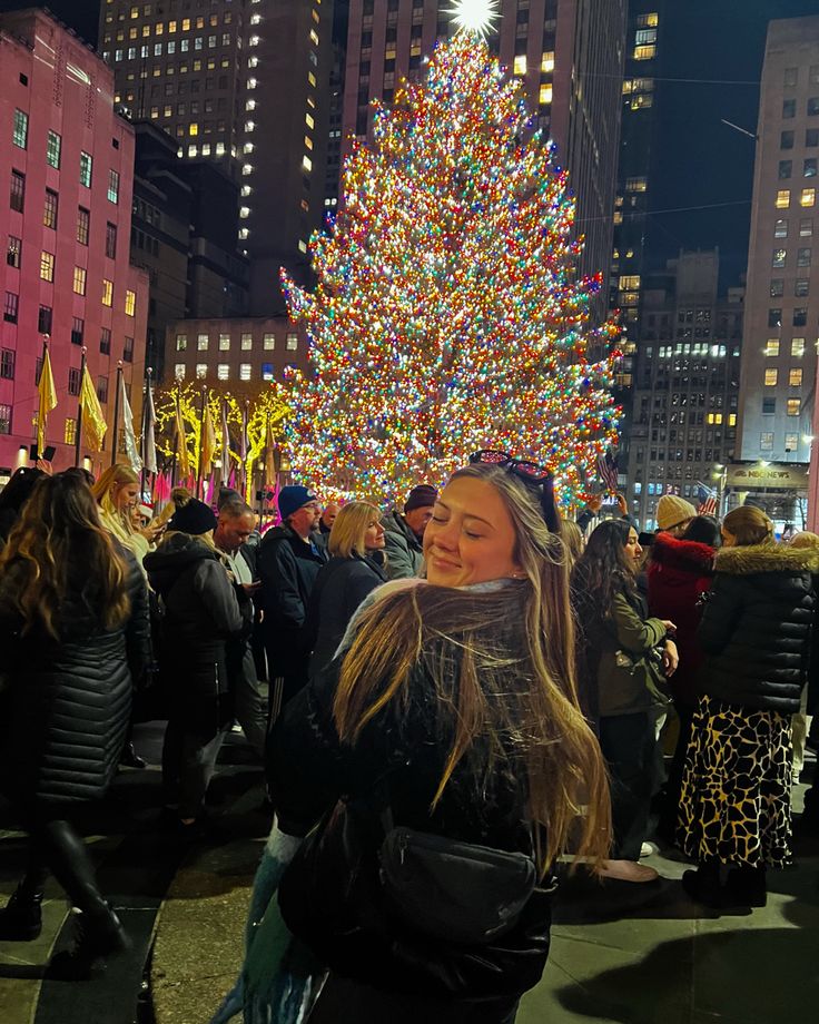 a woman standing in front of a large christmas tree at night with lights on it