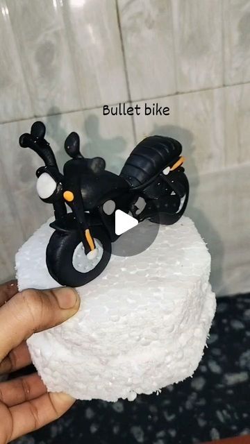 a hand holding a small motorcycle cake on top of a white frosted cake stand