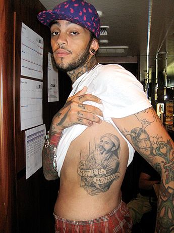 a man with tattoos standing next to a wall in a room filled with posters and papers