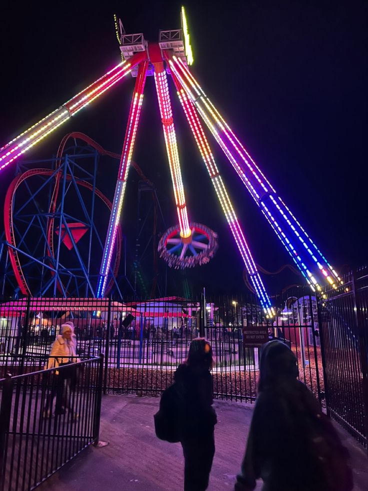 people walking around an amusement park at night with lights on the ferris wheel in the background