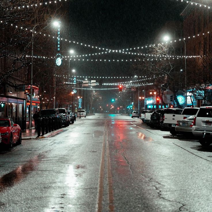 a city street at night with cars parked on both sides and lights strung over the road