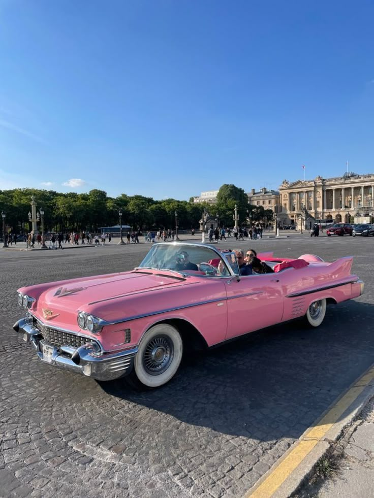 an old pink convertible car parked in front of a building with people walking around it