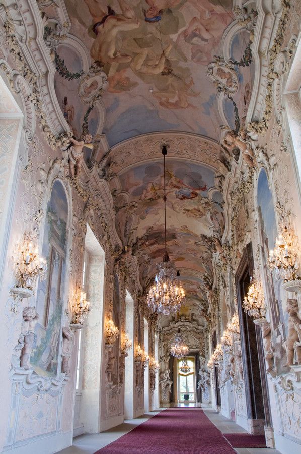 an ornate hallway with chandeliers and paintings on the walls, along with carpeted flooring