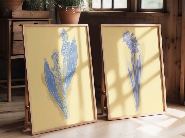 two framed art pieces sitting on top of a hard wood floor next to a potted plant