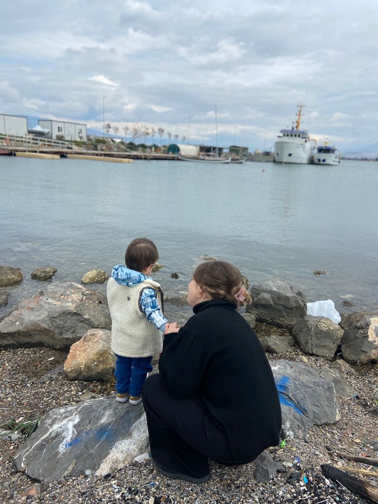 a woman and child are sitting on rocks by the water looking at something in the distance