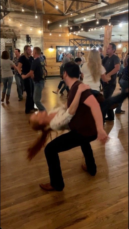 people are dancing in a large room with wooden floors and exposed ceilings, while one person is holding the other's leg