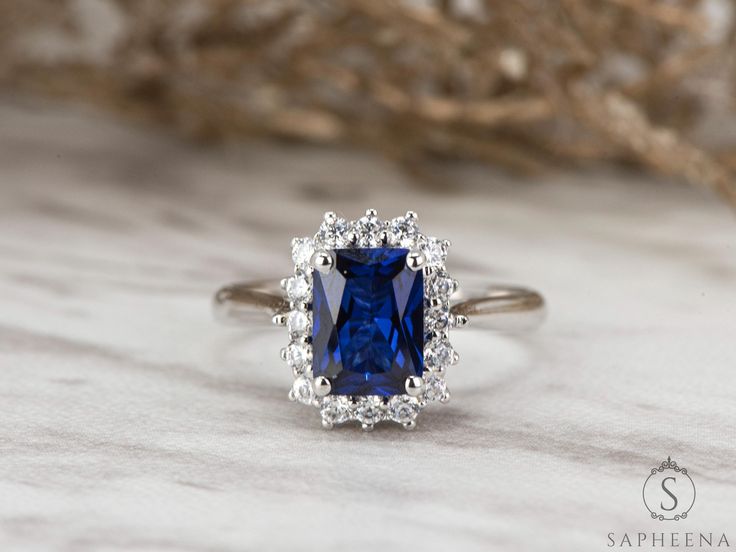 an engagement ring with a blue stone surrounded by white diamonds on a marble surface next to dried twigs