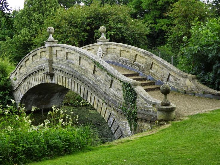 an old stone bridge in the middle of a lush green park with lots of trees