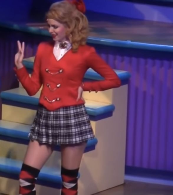 a woman in a red sweater and plaid skirt standing on stage with her hands up