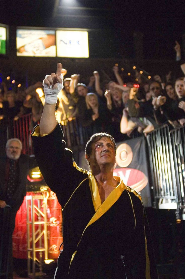 a man in black jacket and yellow scarf holding up his hand with other people behind him