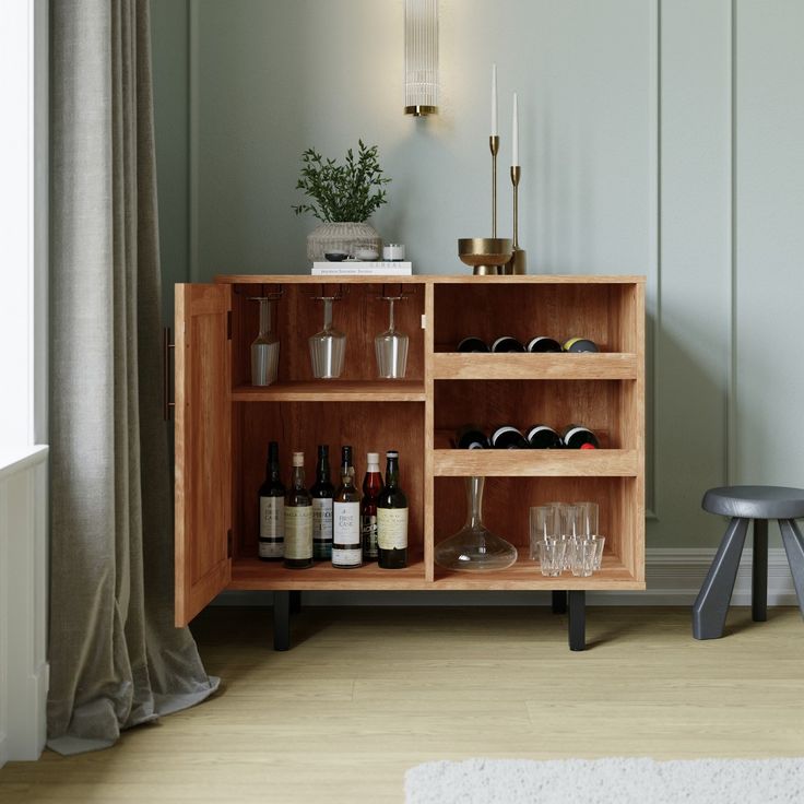 a wooden cabinet with wine glasses and bottles on it next to a stool in front of a window