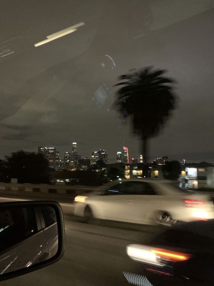 cars driving down the road at night with city lights in the background and palm trees