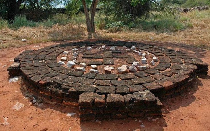 a circular brick fire pit sitting in the middle of a dirt field next to a tree