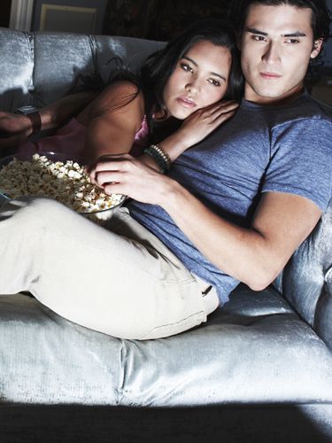 a man and woman sitting on a couch with popcorn in front of their faces as they look at the camera