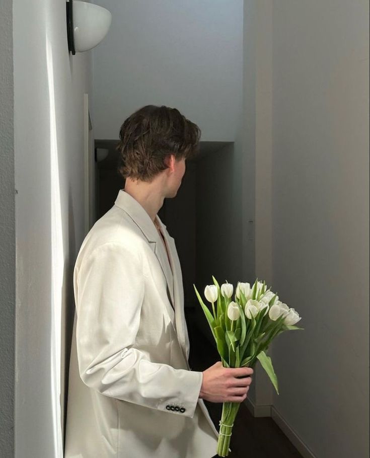 a man in a white suit holding a bouquet of flowers and looking into the mirror