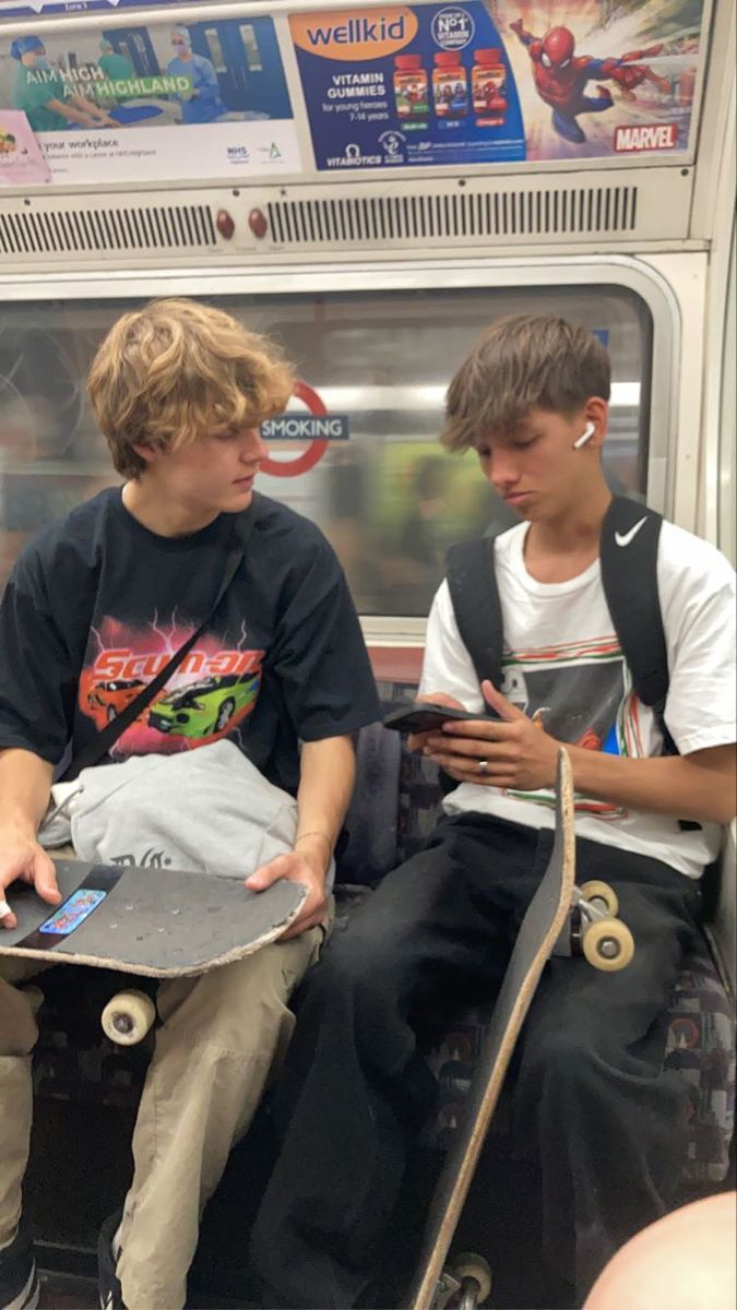 two young boys sitting on a subway looking at their cell phones and holding skateboards