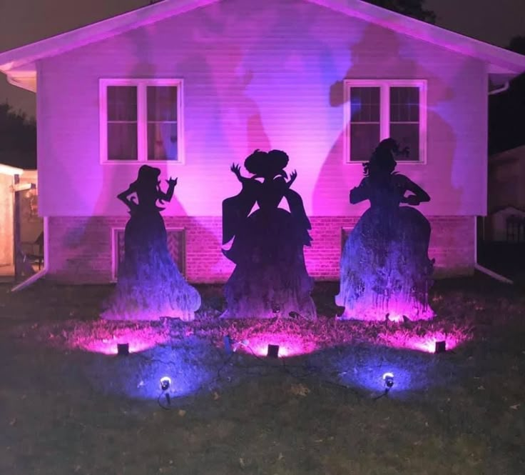 three halloween decorations in front of a house with purple lights on the lawn and two people standing next to each other