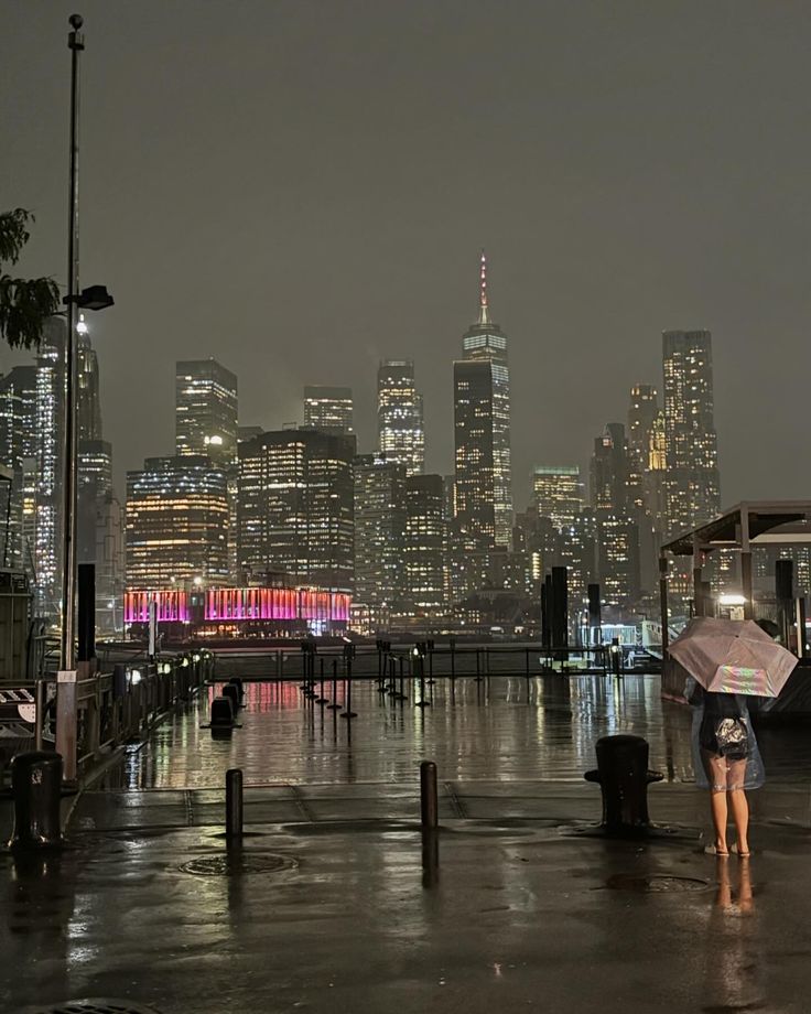 a woman holding an umbrella standing in the rain next to a body of water at night