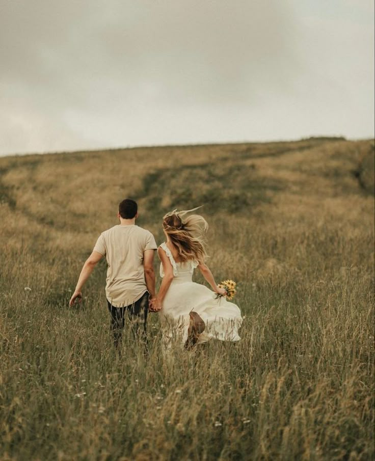 a man and woman walking through tall grass in the middle of an open field, holding hands