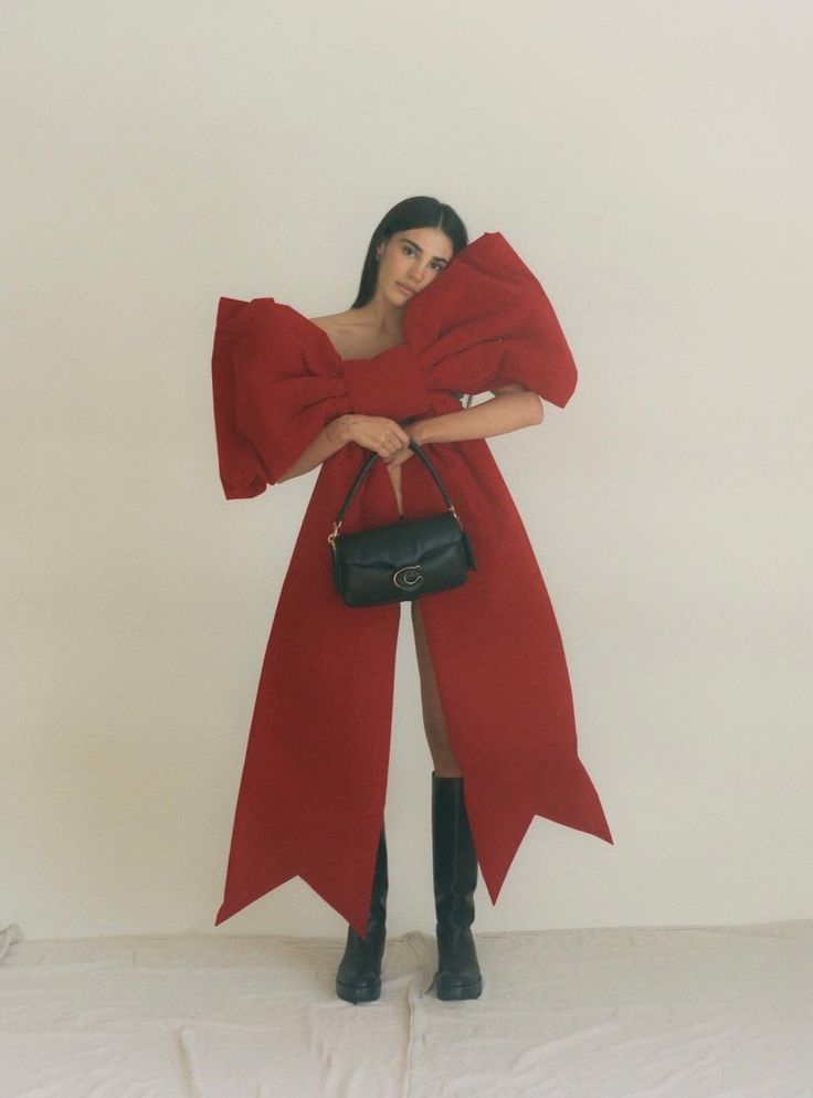 a woman wearing a red coat and holding a black handbag in front of a white wall