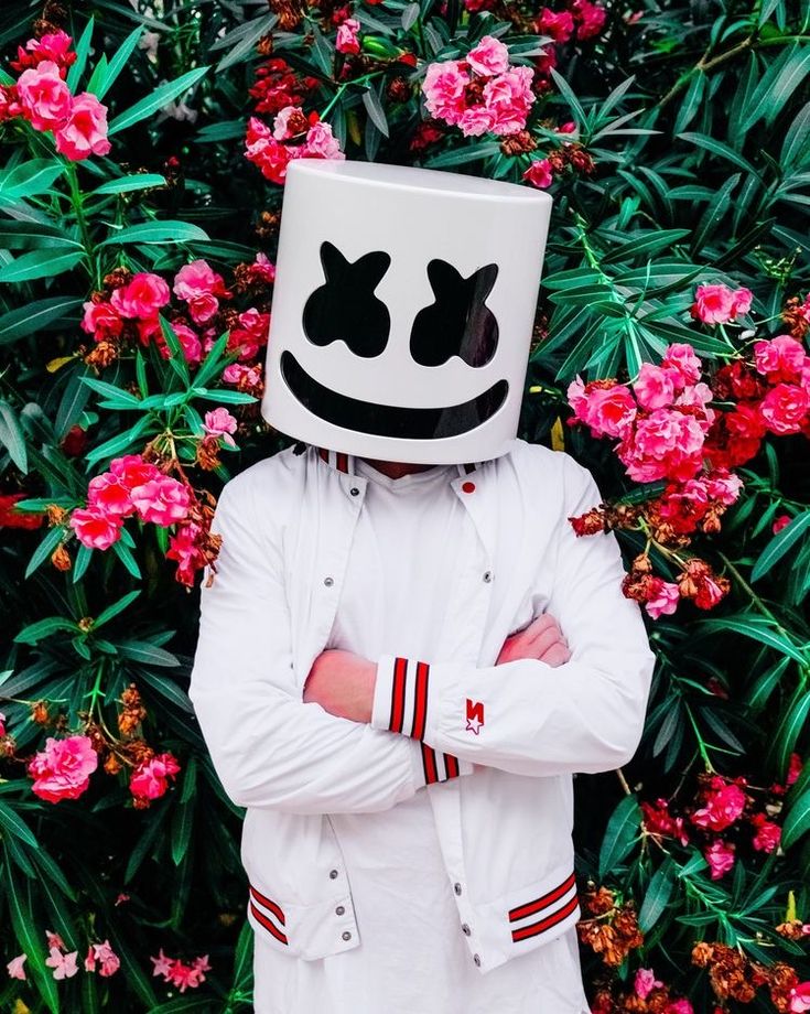 a person wearing a marshmallow mask standing in front of pink flowers with their arms crossed