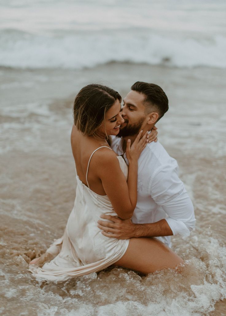 a man and woman are sitting in the water at the beach, hugging each other