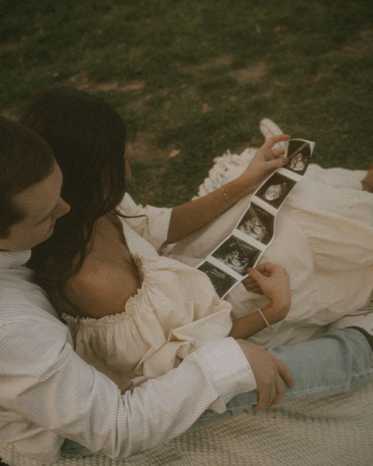 a man and woman are sitting on the grass together, holding an album in their hands