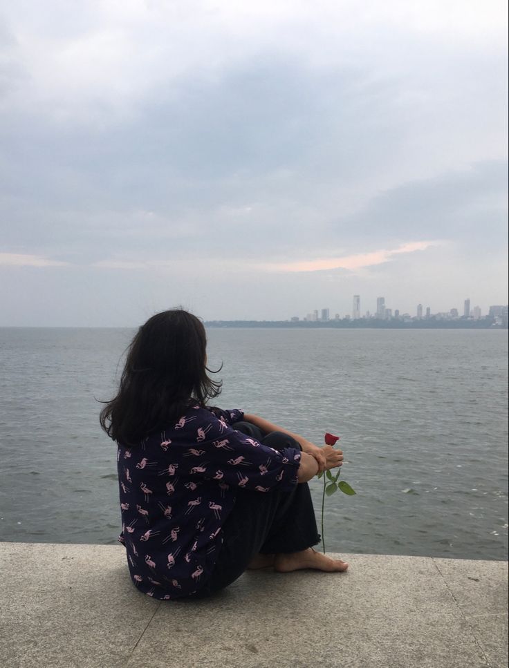 a woman sitting on the edge of a pier looking out at the water with a rose in her hand