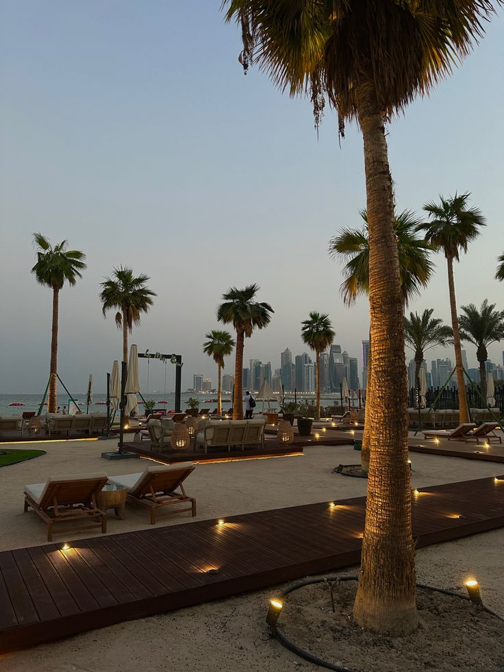 palm trees and lounge chairs are lit up by lights on the boardwalk in front of a cityscape