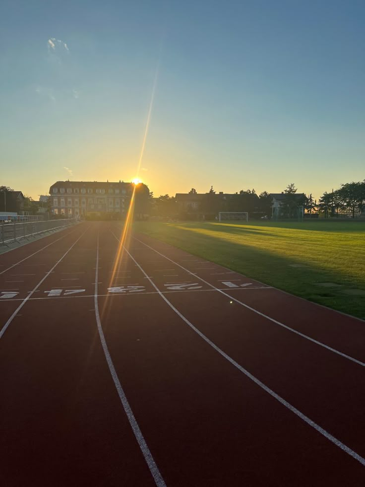 the sun is setting over a running track in an empty area with grass and buildings