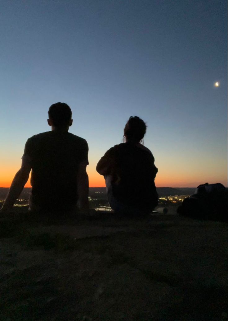 two people sitting on top of a hill watching the sunset