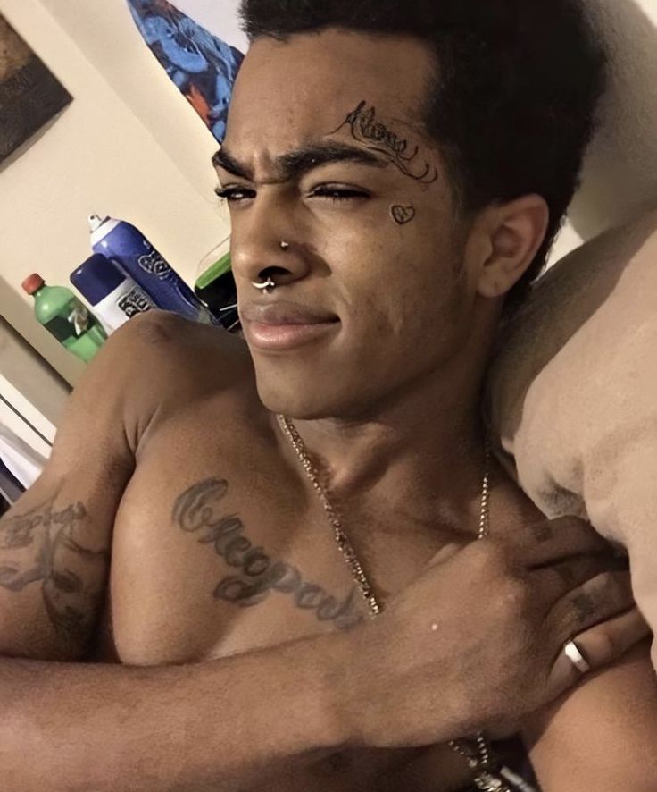 a shirtless man with tattoos on his chest sitting in front of a bed and looking at the camera