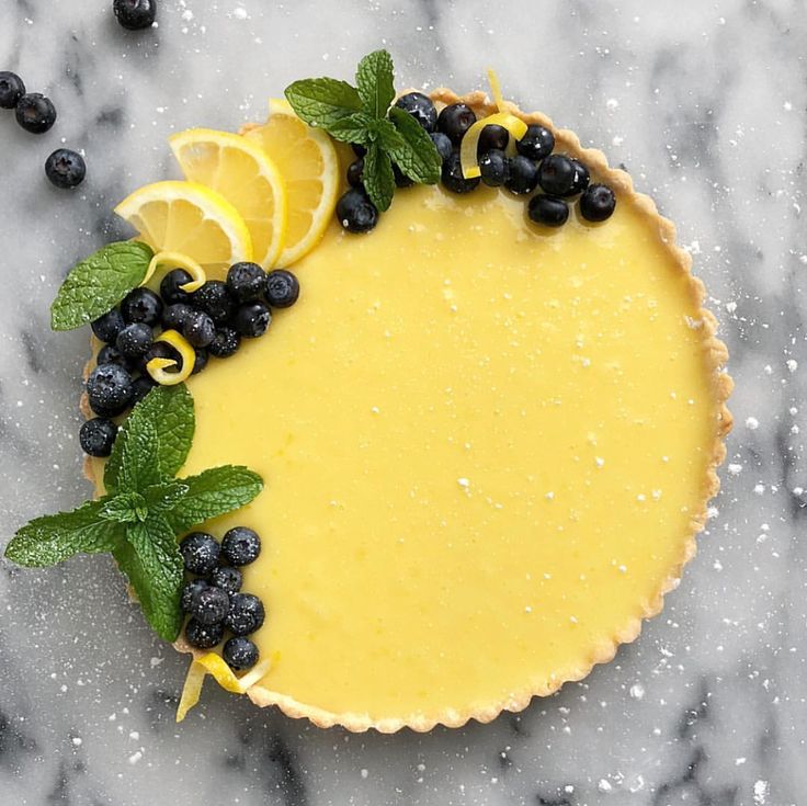 a lemon tart topped with blueberries and mint leaves on a marble counter top