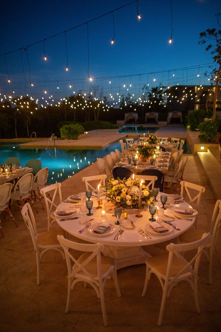 an outdoor dining area next to a pool at night with lit candles on the tables