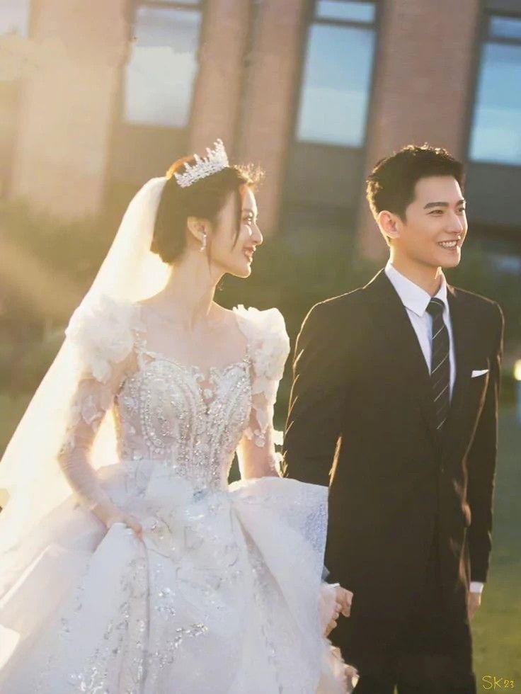 a man and woman dressed in formal wear walking together on the grass with buildings in the background