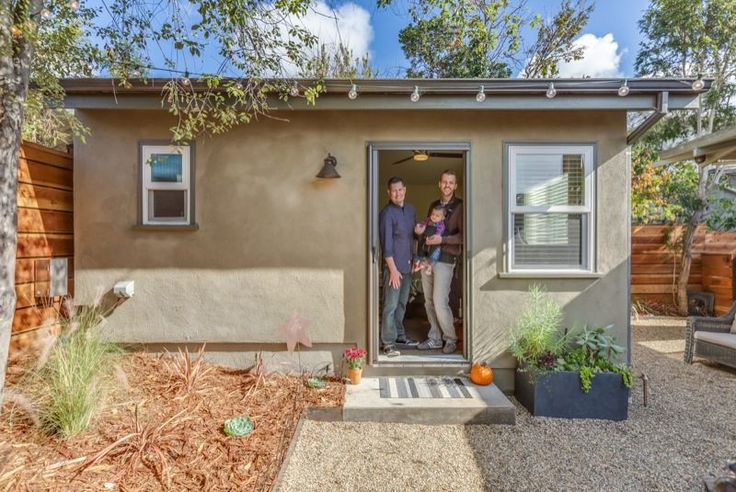 two people are standing in the doorway of a small house with plants and trees around it