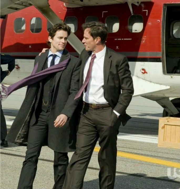 two men in suits walking towards an airplane with a purple scarf around their neck and one man wearing a suit