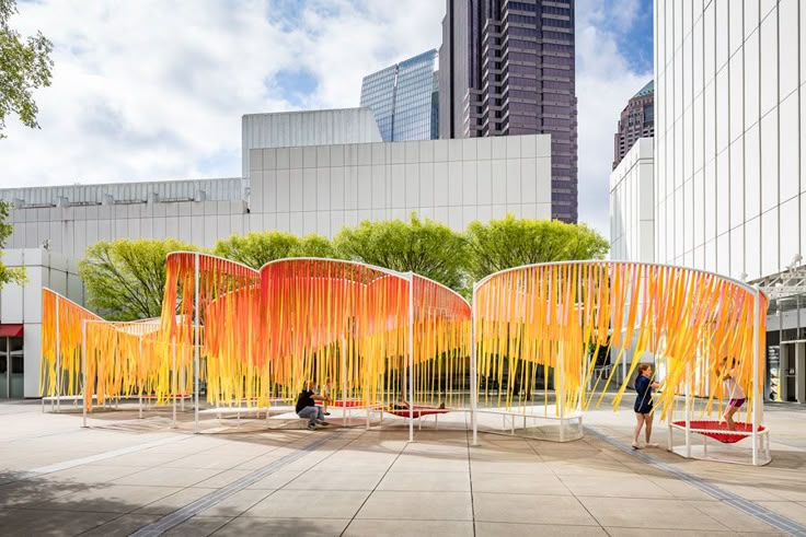 two people are standing in front of an orange sculpture on the ground near tall buildings