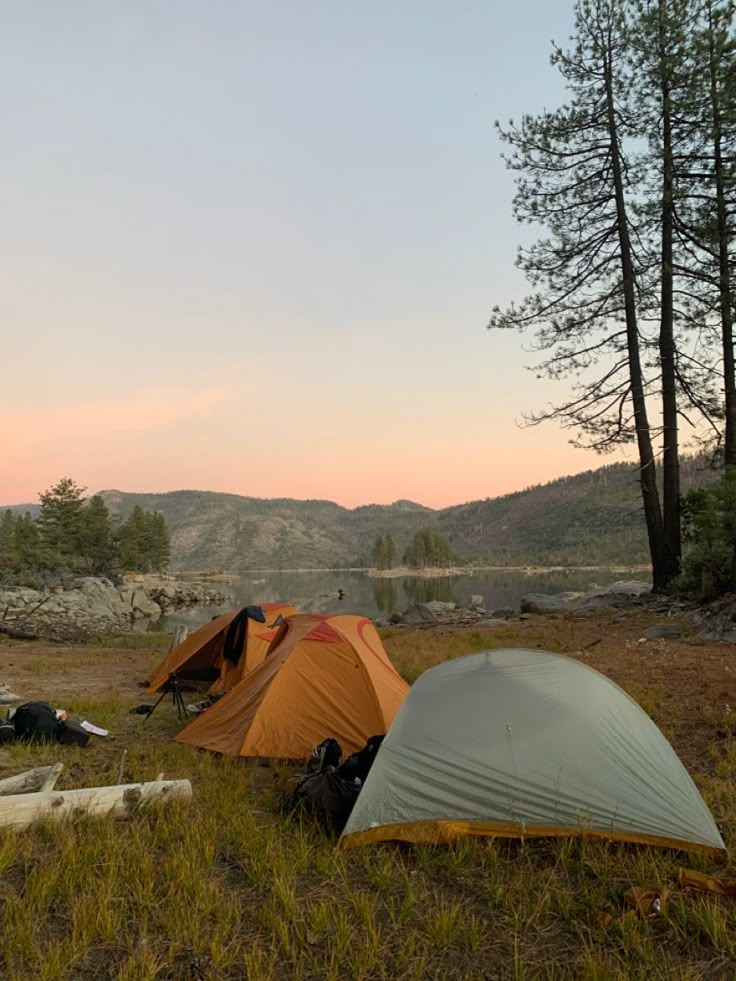 two tents are pitched up in the grass near some trees and water at sunset with mountains in the background