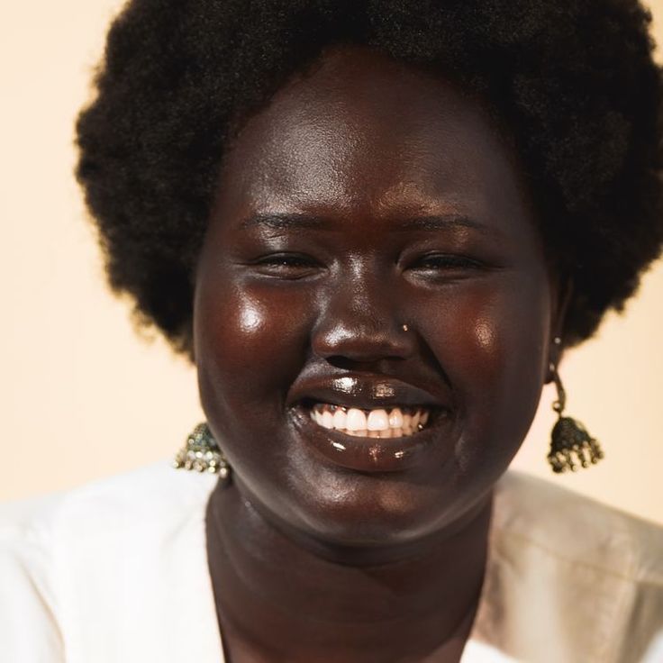 a woman with dark skin and large earrings smiles at the camera while wearing white shirt