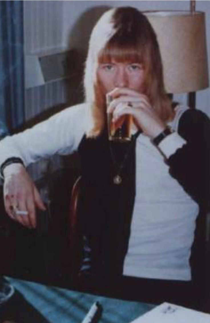 a woman sitting at a table drinking from a glass in front of a lamp and papers