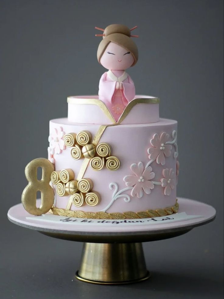 a pink and gold birthday cake with a doll on it's top, sitting on a plate