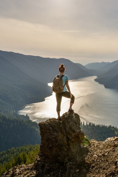 a woman standing on top of a cliff with a backpack looking at the water and mountains