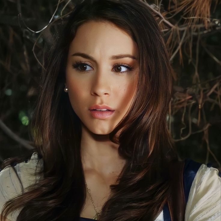 a beautiful young woman with long brown hair and blue eyes posing for a photo in front of a tree