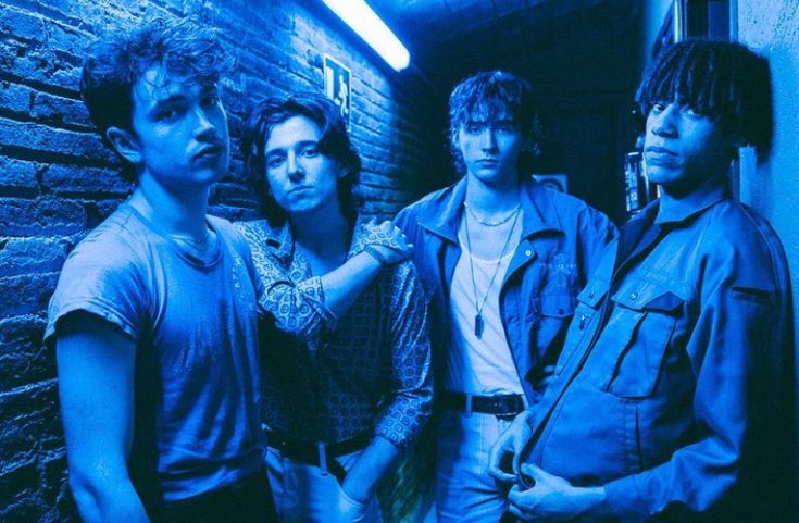 four young men standing next to each other in front of a brick wall and blue light