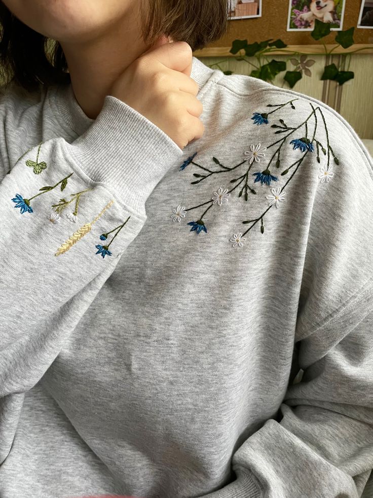 a woman wearing a gray sweatshirt with blue flowers on the front and side, holding her hand to her chest